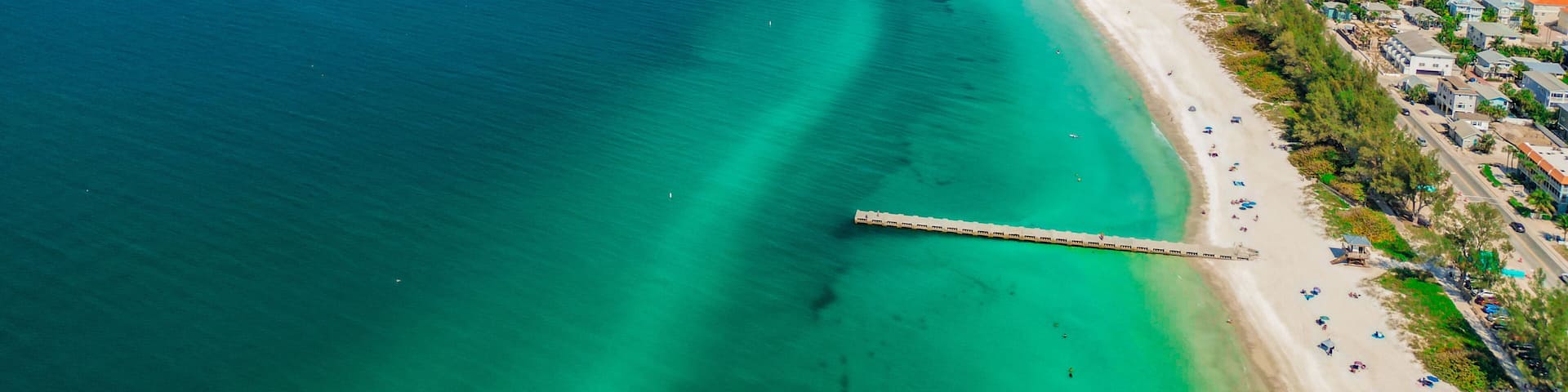 A coastal town with a long pier extending into the turquoise sea, a sandy beach, and residential buildings along the shore.
