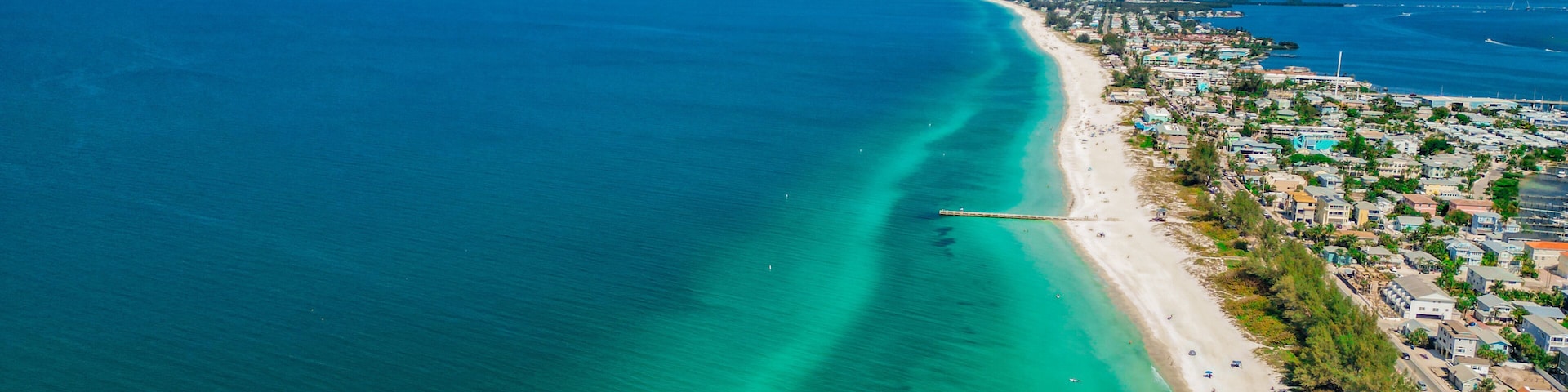 A coastal town with a long pier extending into the turquoise sea, a sandy beach, and residential buildings along the shore.