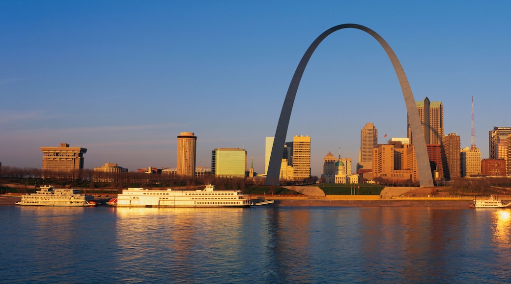 This is the skyline at sunrise. It is situated along the Mississippi River. There are riverboats on the water with the St. Louis Arch in clear view.