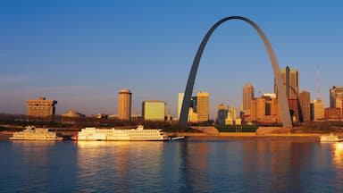 This is the skyline at sunrise. It is situated along the Mississippi River. There are riverboats on the water with the St. Louis Arch in clear view.
