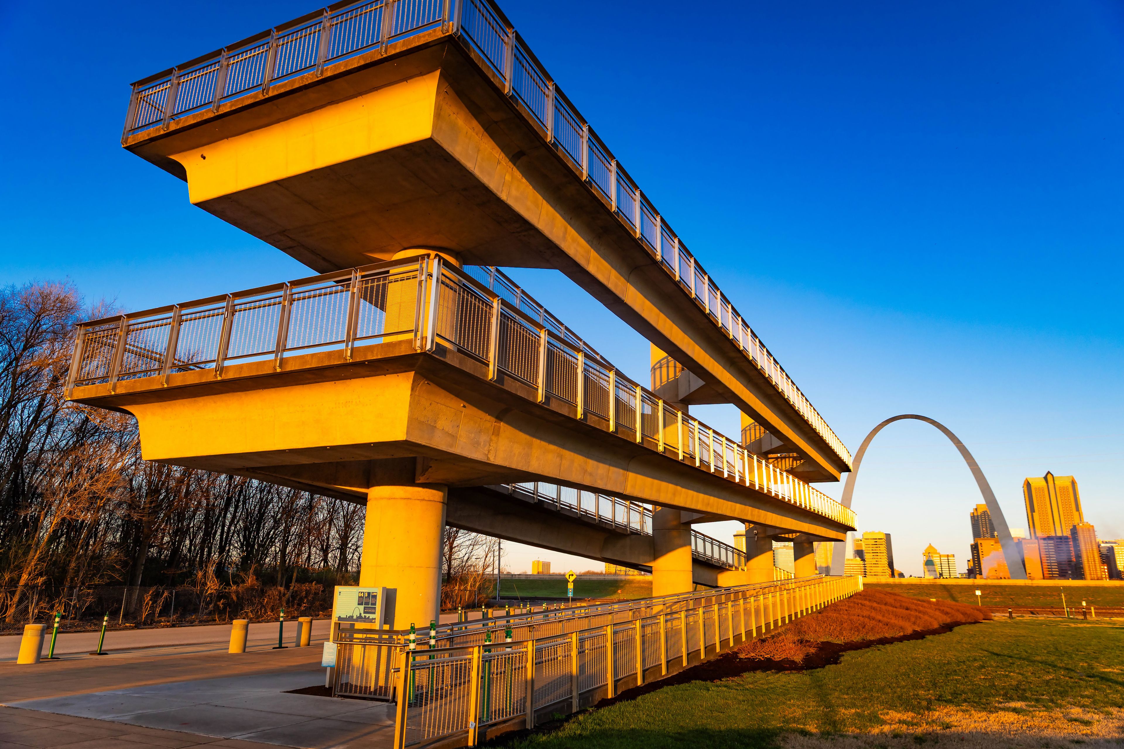 A multi-level pedestrian bridge with railings, a clear blue sky, and a city skyline in the background.