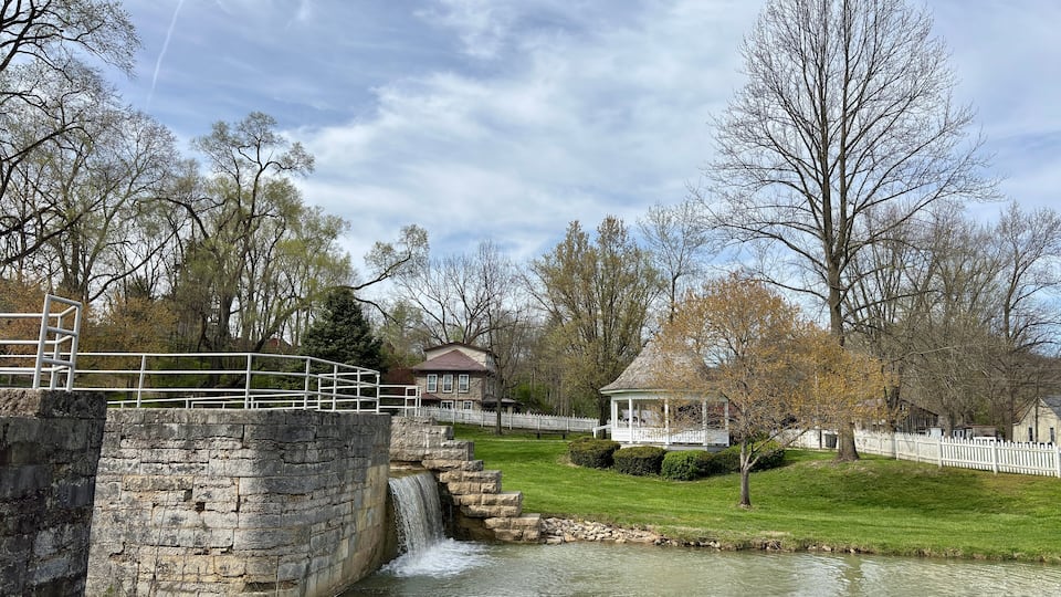 A stone bridge, a small waterfall, a gazebo, and a white picket fence in a park.