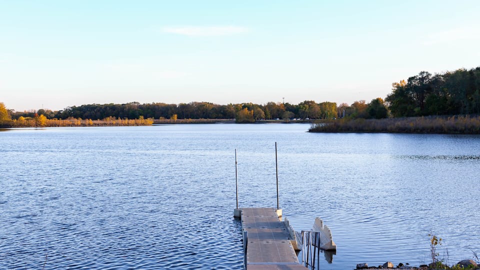 A calm lake with a wooden dock extending into the water, surrounded by trees and a clear sky.