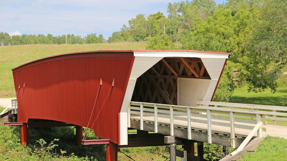 A red covered bridge with a white roof and wooden deck, spanning a small stream in a rural area with green fields and trees in the background.