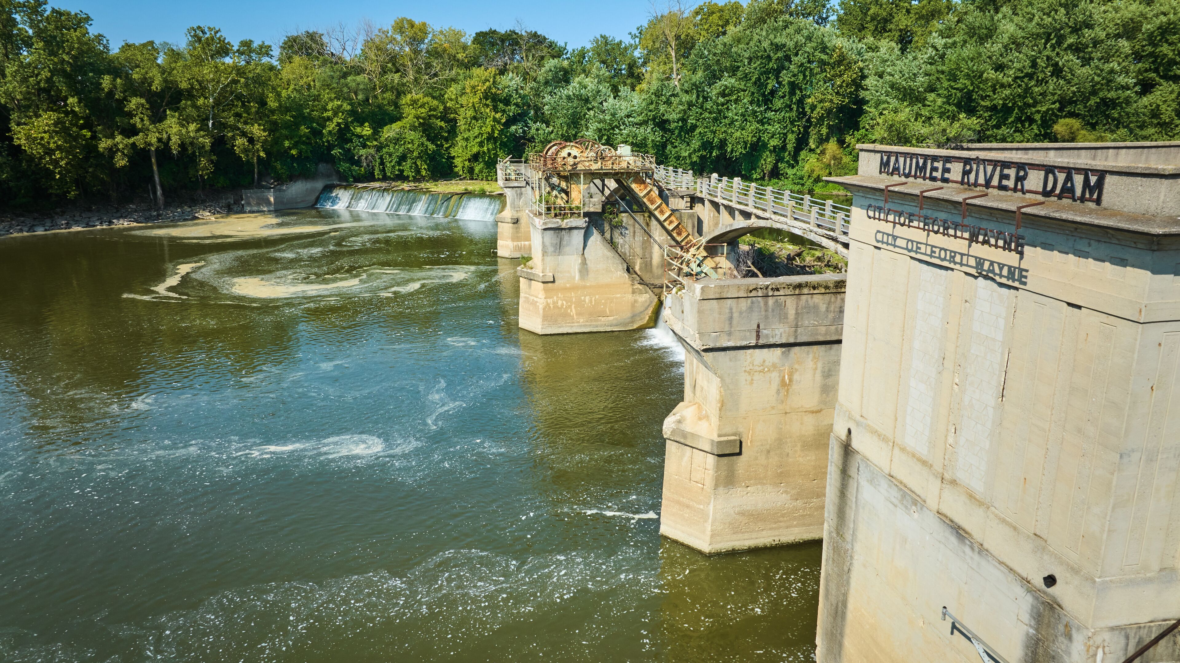 A dam with a bridge, water flow, and surrounding greenery.