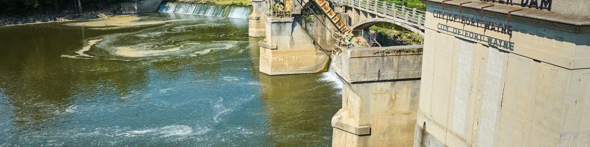 A dam with a bridge, water flow, and surrounding greenery.