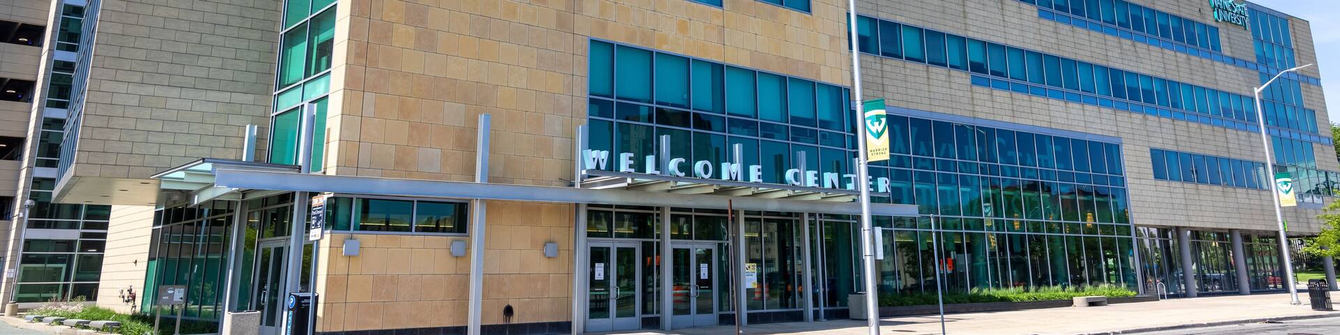 A modern building with 'Wayne State University' signage, a 'Welcome Center' entrance, and a clear blue sky.
