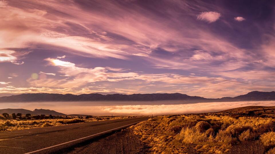 A road leading through a dry, grassy landscape with mountains in the background and a dramatic sky with clouds.