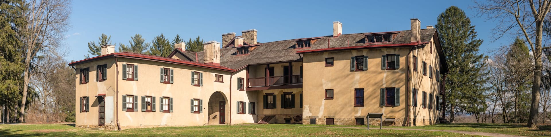 A large, historic building with multiple chimneys and a red roof, surrounded by a grassy area and trees.
