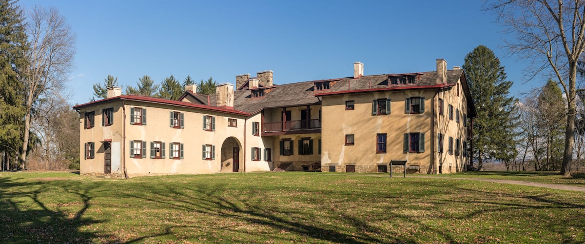 A large, historic building with multiple chimneys and a red roof, surrounded by a grassy area and trees.