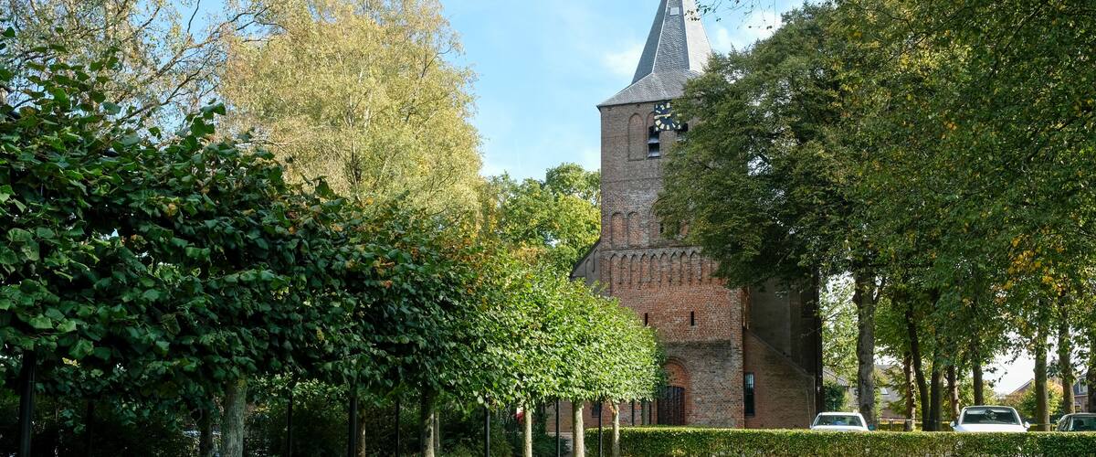 A historic church with a tall tower and a clock, surrounded by lush greenery and trees.