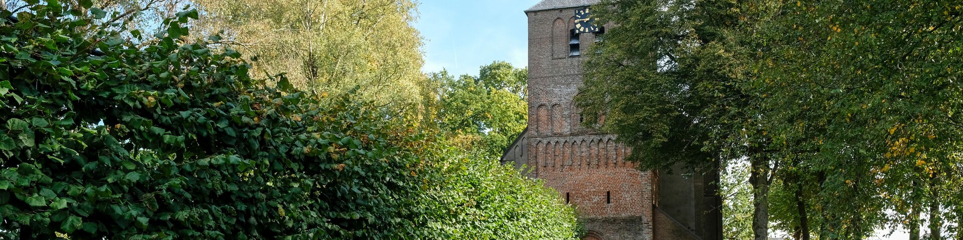A historic church with a tall tower and a clock, surrounded by lush greenery and trees.