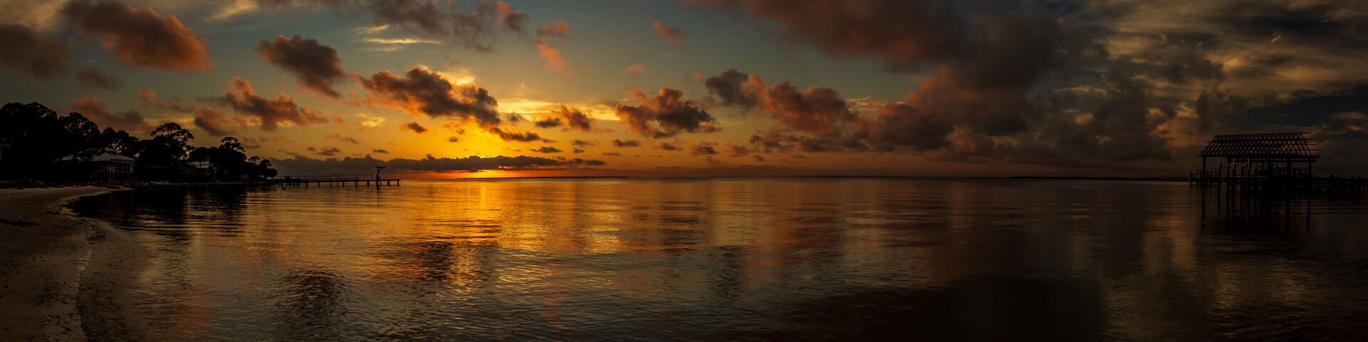 A sunset over a calm body of water with a pier on the right and a beach on the left.