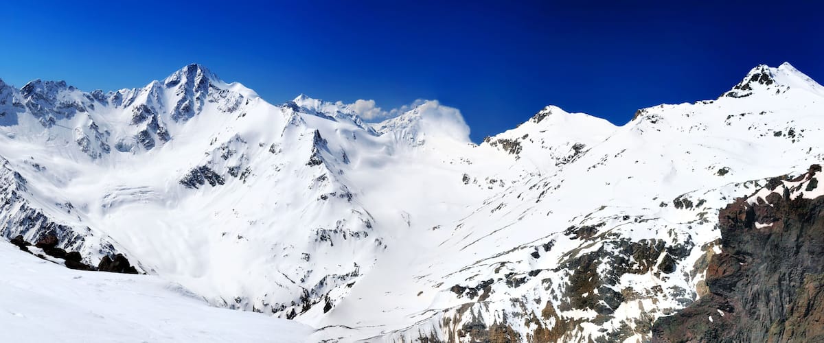 A snow-covered mountain range with sharp peaks and rocky outcrops under a clear blue sky.