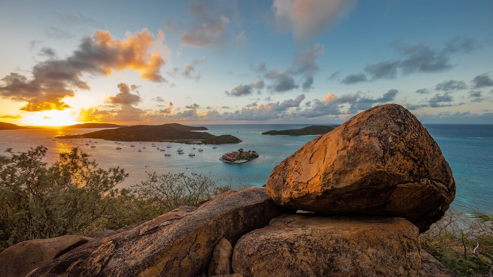 A sunset over a coastal area with a large rock formation in the foreground and several boats on the water.