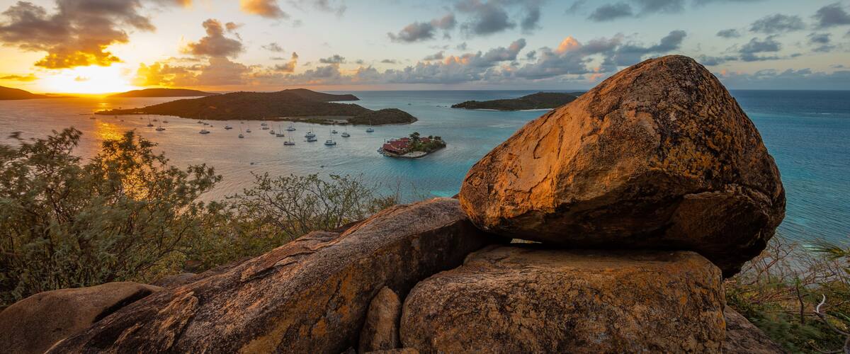 A sunset over a coastal area with a large rock formation in the foreground and several boats on the water.