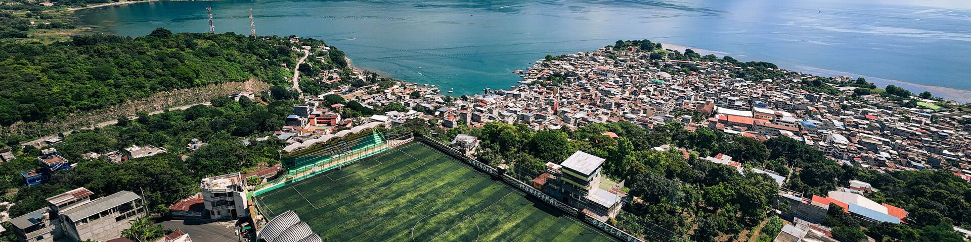 Aerial view of a soccer field nestled between lush greenery and the tranquil waters of Lake Atitlan, with homes cascading down the hillside, Antigua Guatemala, Sacatepequez Department, Guatemala.