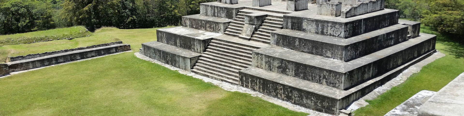Uma grande estrutura piramidal antiga com uma escadaria que leva ao topo, cercada por grama verde exuberante e árvores.