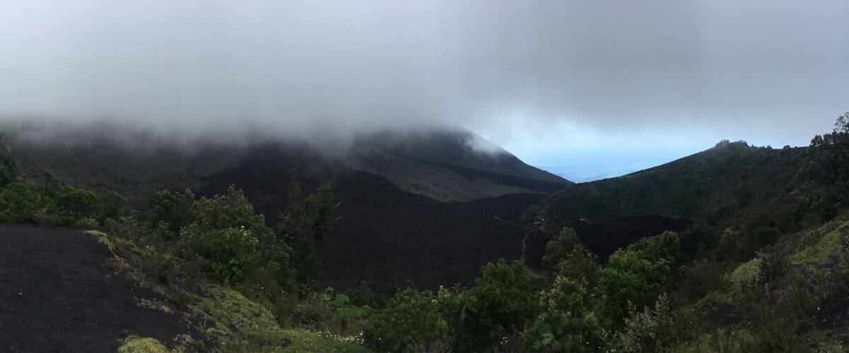 A volcanic landscape with dense vegetation and a mountain partially obscured by clouds.