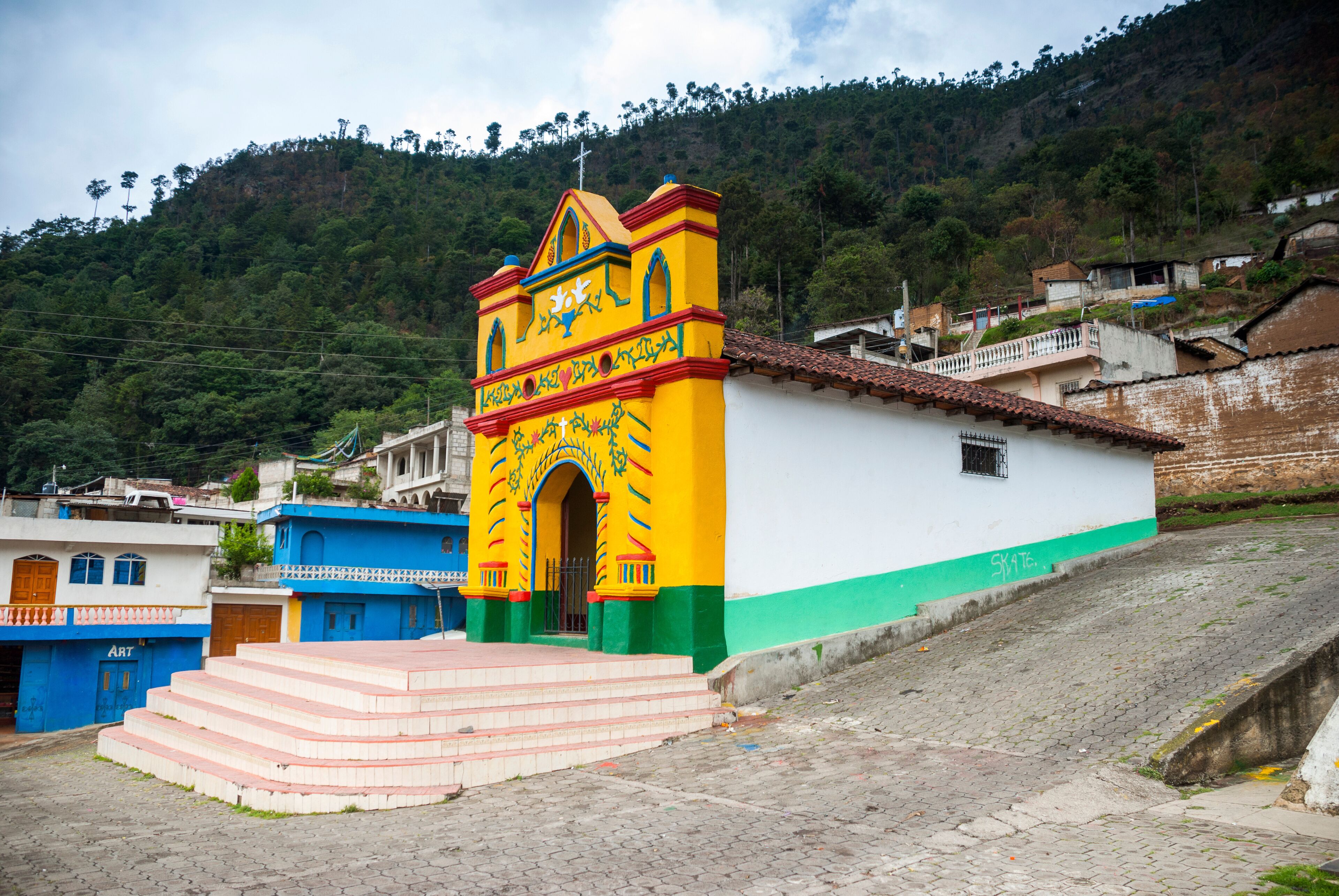 A colorful church with a bell tower, steps leading up to it, and a hillside in the background.