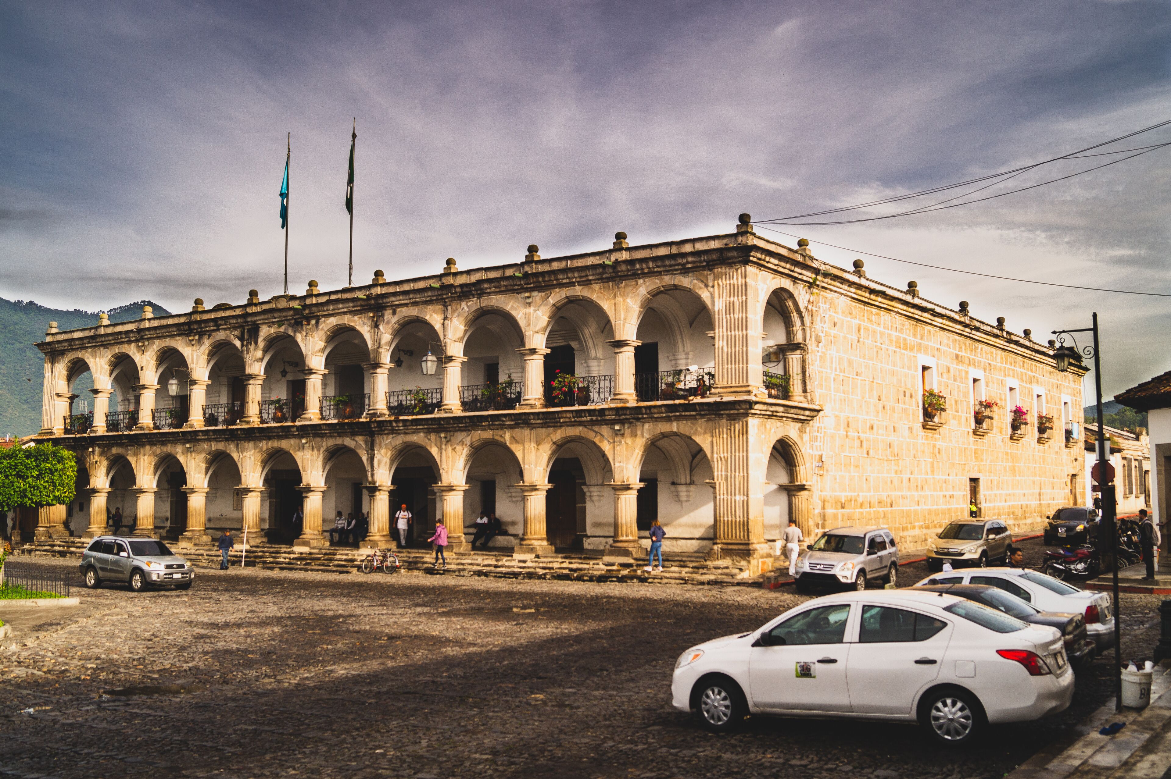A historic building with multiple arches and columns, surrounded by parked cars and people.