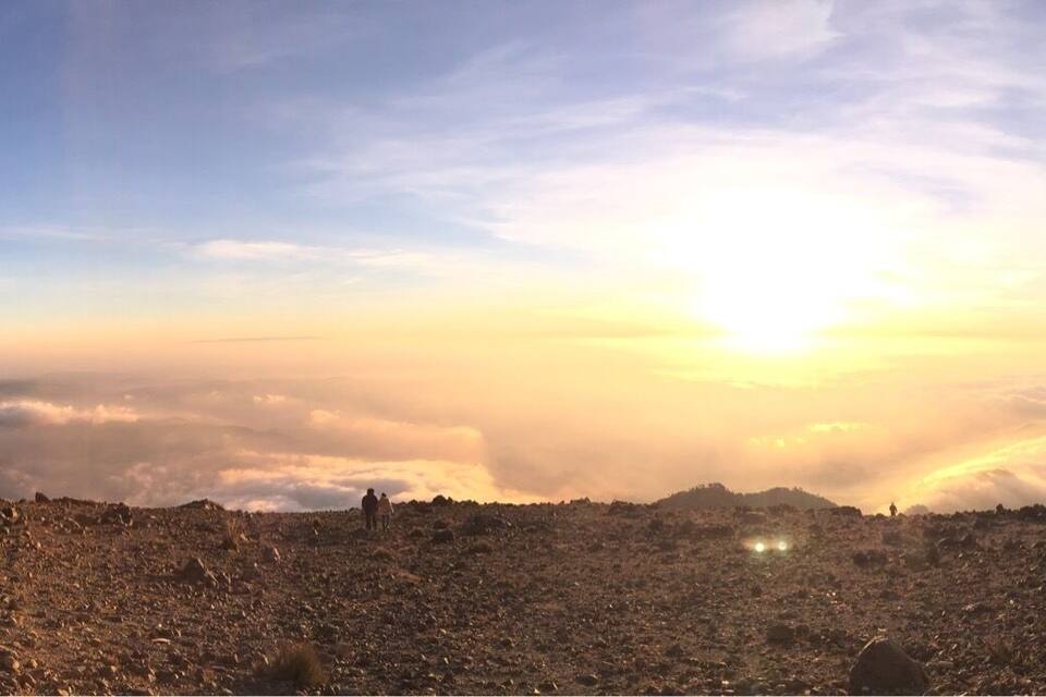 Paisagem montanhosa com nascer ou pôr do sol, terreno rochoso e um grupo de pessoas em um cume.