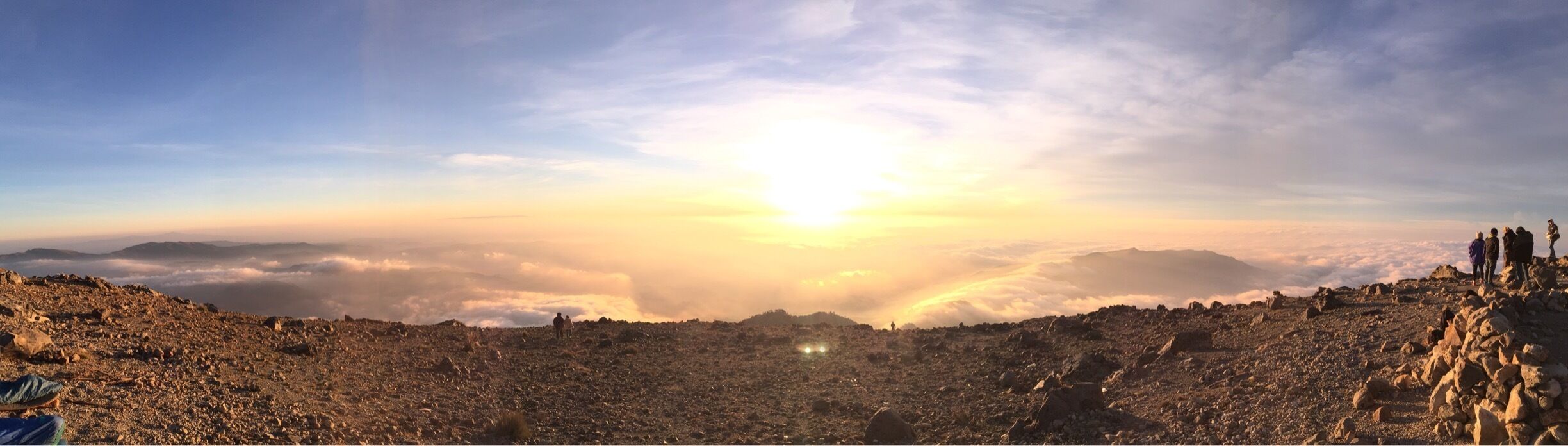 A mountainous landscape with a sunrise or sunset, rocky terrain, and a group of people standing on a ridge.