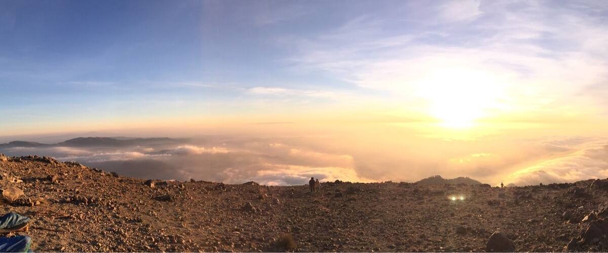 A mountainous landscape with a sunrise or sunset, rocky terrain, and a group of people standing on a ridge.