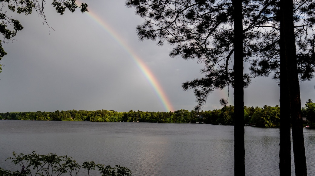 Un lac où se reflète un arc-en-ciel, entouré d’arbres et surplombé d’un ciel nuageux.