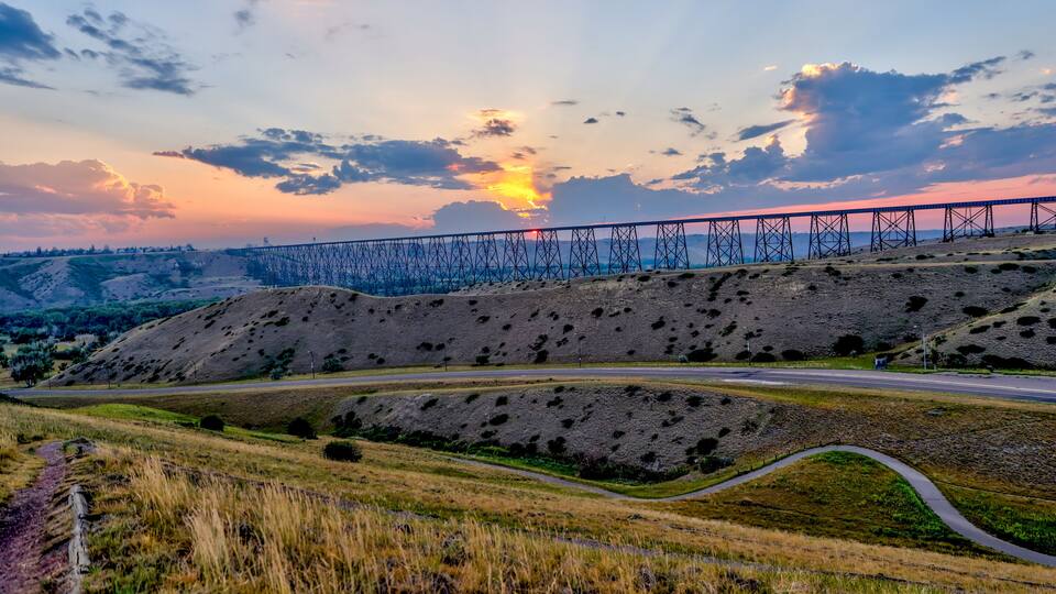 A sunset over a bridge, a winding road, and a hilly landscape.