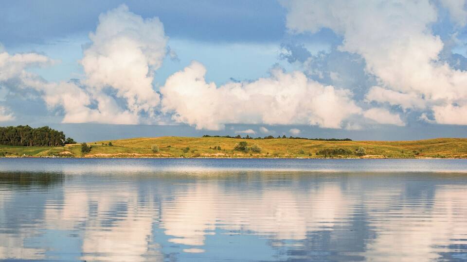 A calm lake with a grassy hill in the background and a partly cloudy sky.