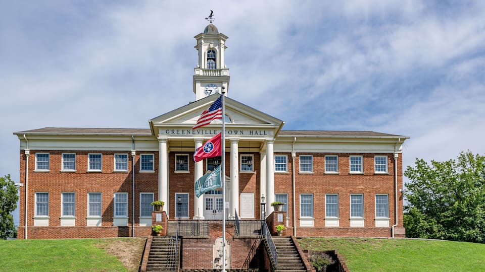 A brick building with a central tower, a flagpole with flags, and a sign reading 'Greenville Town Hall'.