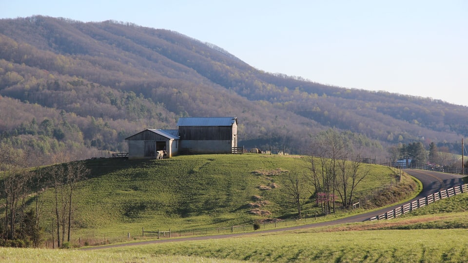 A rural landscape with a barn on a hill, a winding road, and forested hills in the background.