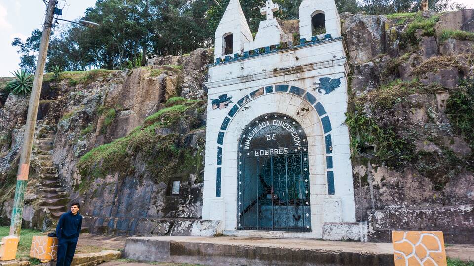 A white stone structure with a cross on top, a gate with the inscription "IMMACULADA CONCEPCION DE LOURDES", and a monkey on the rock behind it.