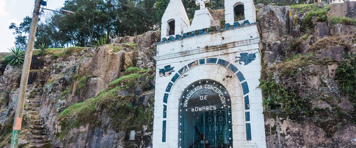 A white stone structure with a cross on top, a gate with the inscription "IMMACULADA CONCEPCION DE LOURDES", and a monkey on the rock behind it.