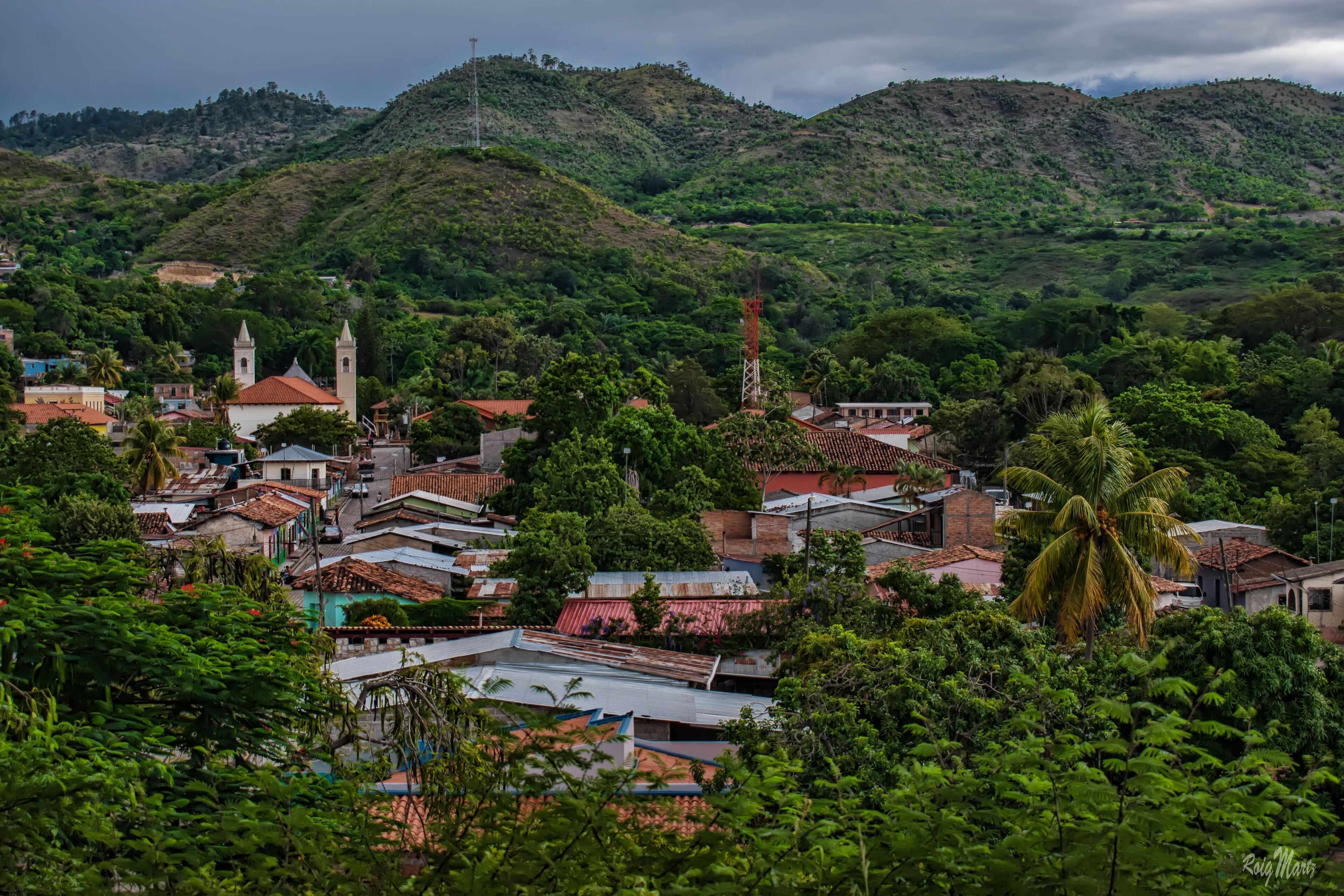 A town nestled in a valley with hills in the background, featuring a church with two towers and a prominent tower structure.
