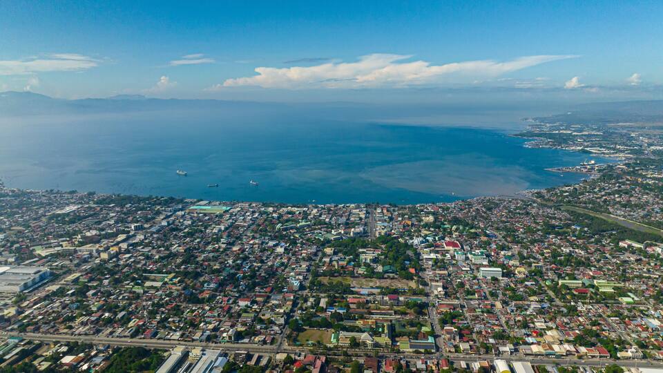 An aerial view of a coastal city with dense urban development, a large body of water, and a distant mountain range.