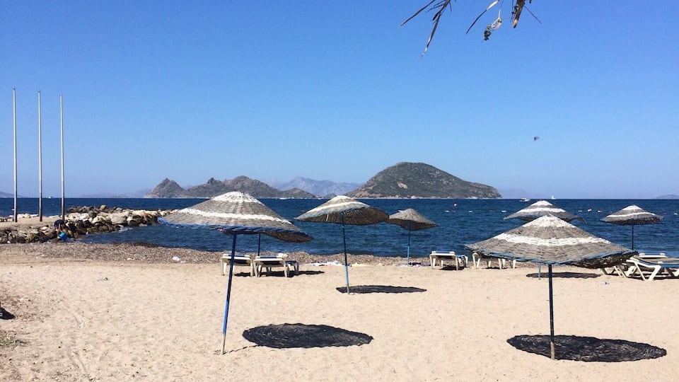 Una playa con sombrillas y sillas de descanso, una costa rocosa y montañas al fondo.