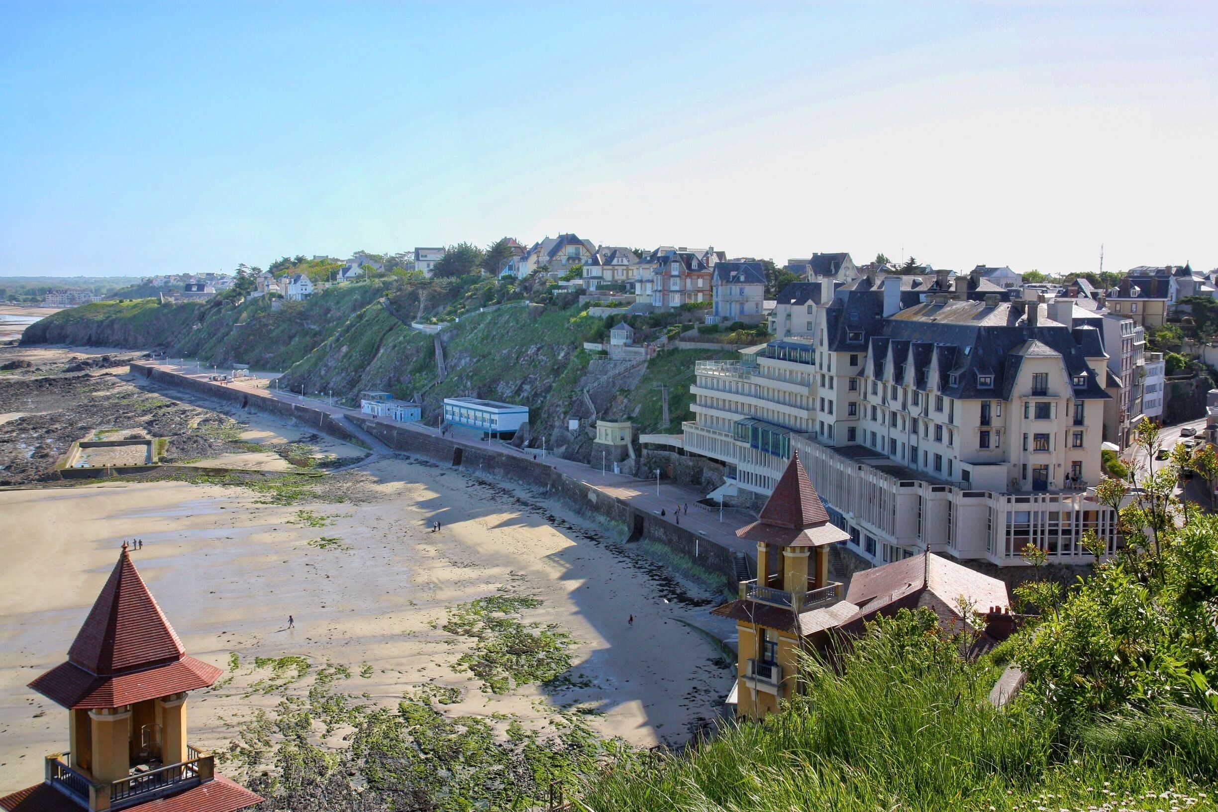 A coastal town with a sandy beach, a lighthouse, and residential buildings on a hillside.