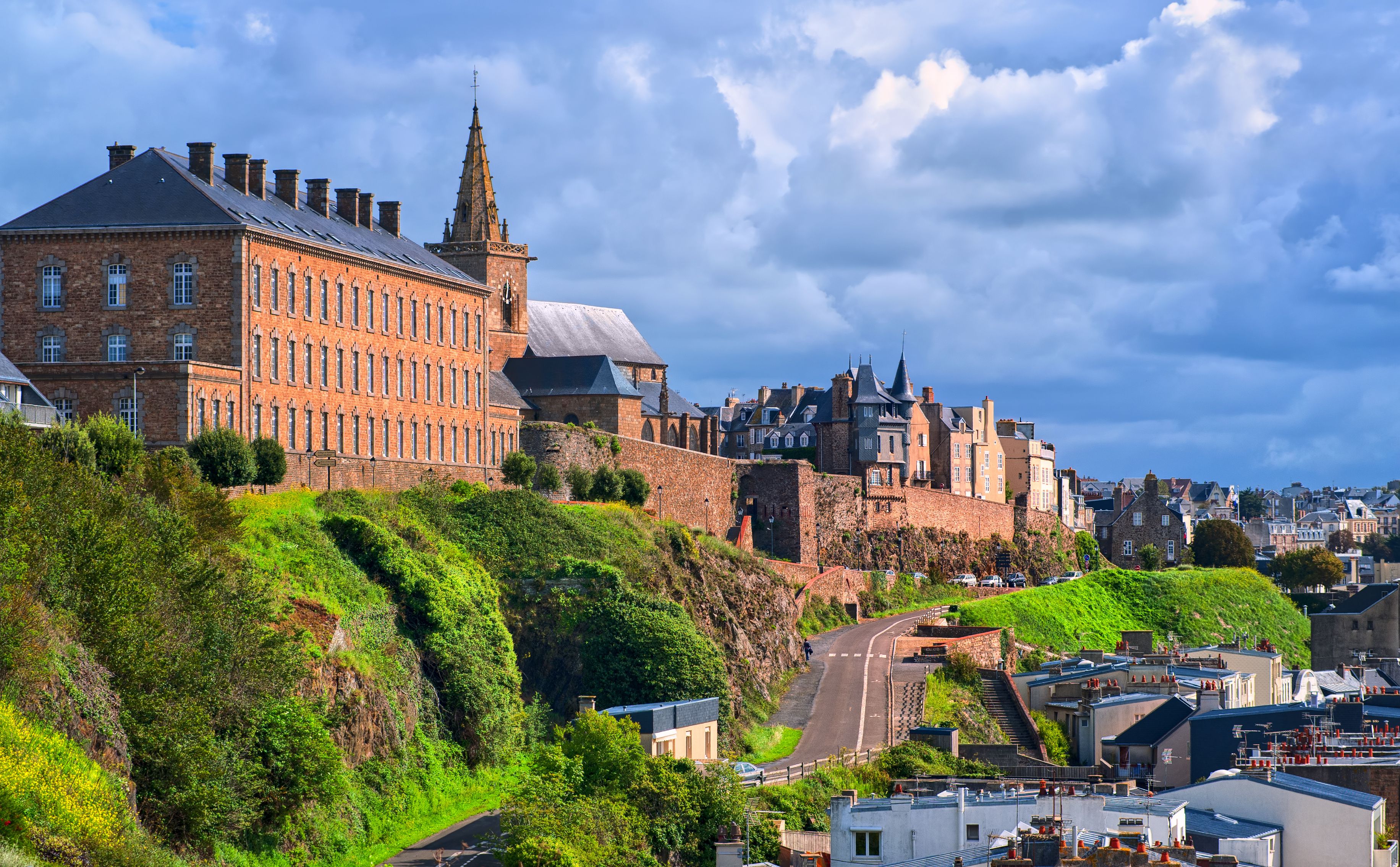 A historic building with a prominent tower, a steep hill with greenery, and a winding road descending the hill.