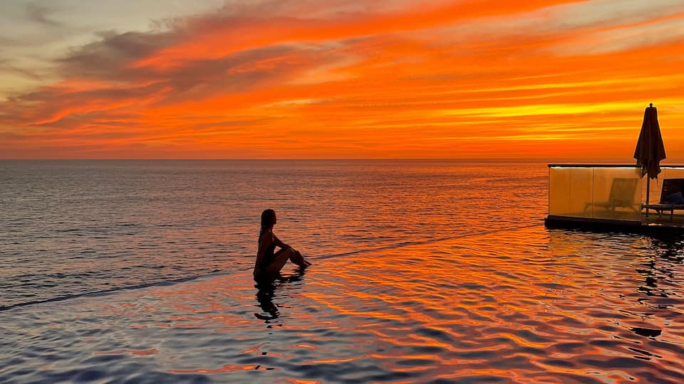 A sunset over a calm sea with a person sitting in the water and a structure with a canopy on the right.
