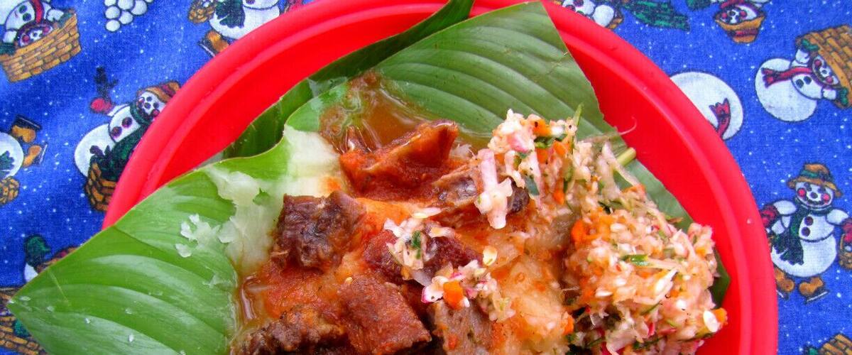 A meal served on a banana leaf with a red bowl, including meat, rice, and a side of coleslaw.