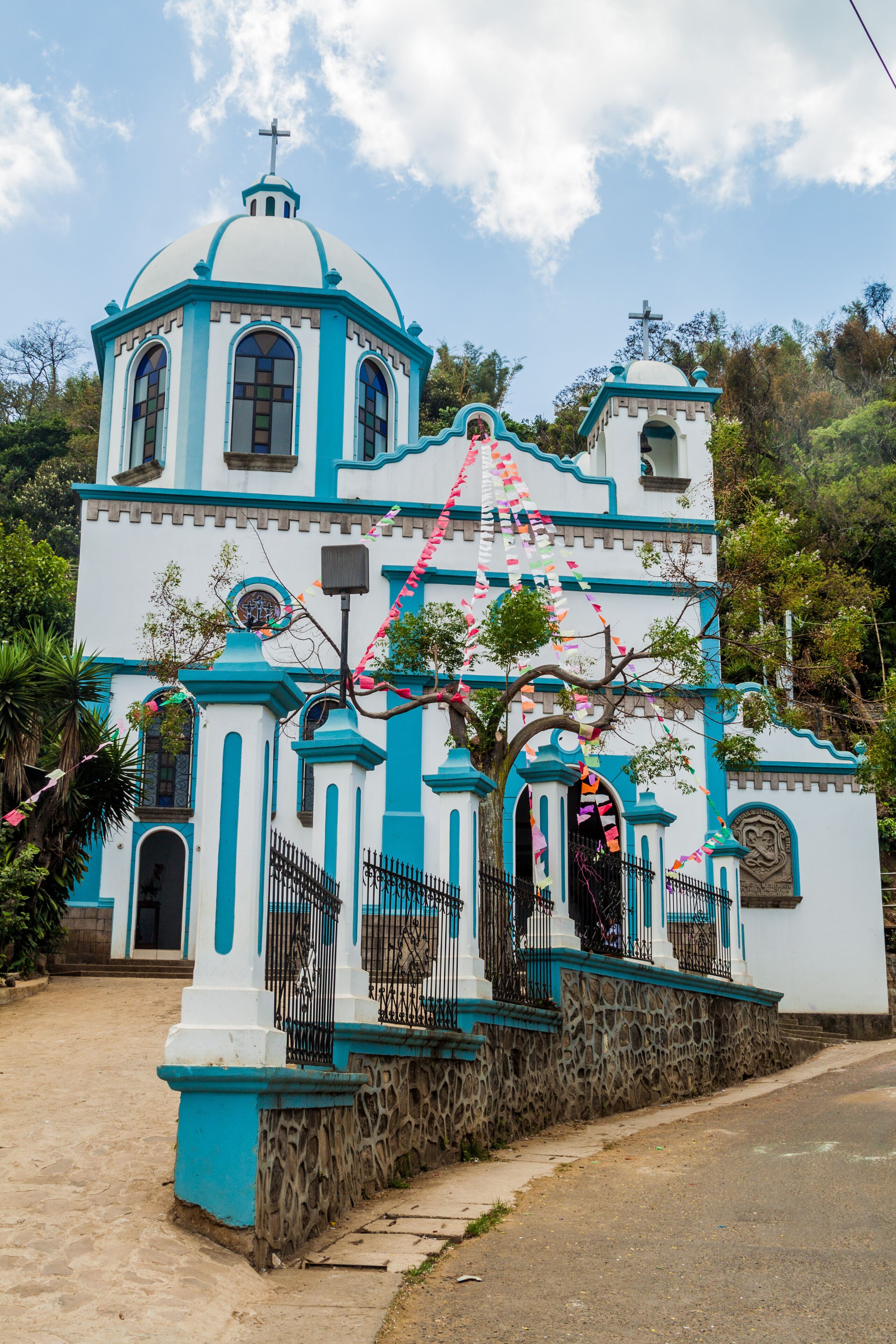 A white and blue church with a cross on top, surrounded by trees and a stone wall.
