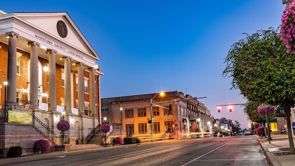 A historic courthouse with columns and a clock tower, a street with traffic lights, and flowering trees lining the sidewalk.