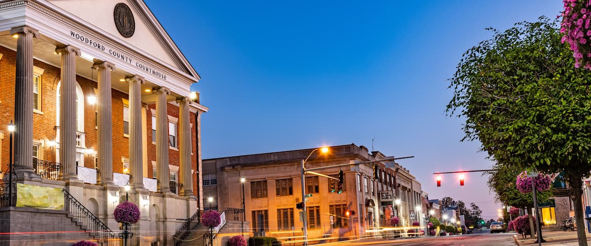 A historic courthouse with columns and a clock tower, a street with traffic lights, and flowering trees lining the sidewalk.