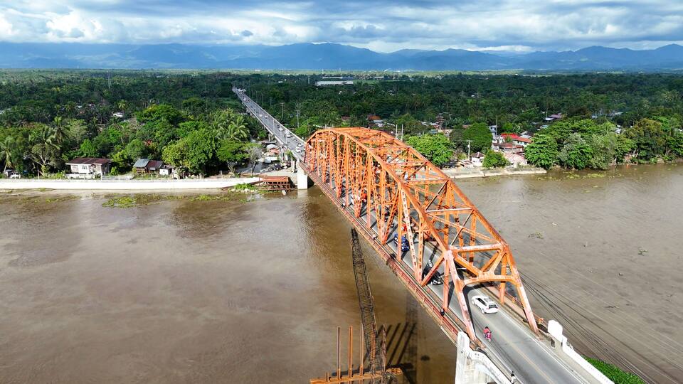 A large orange bridge spanning a river with a backdrop of green hills and a cloudy sky.