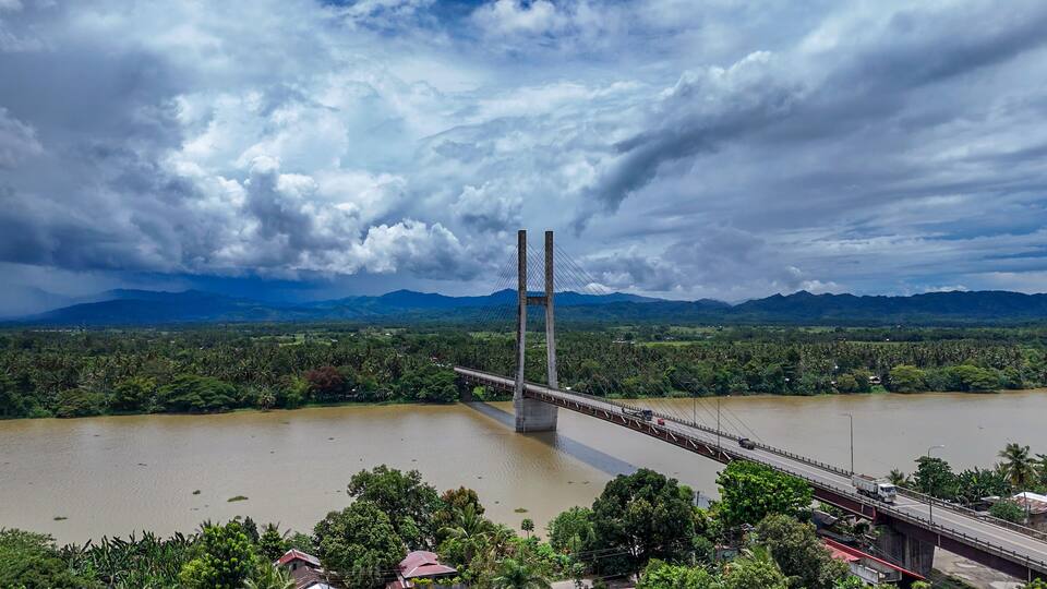A suspension bridge over a river with a mountainous backdrop.