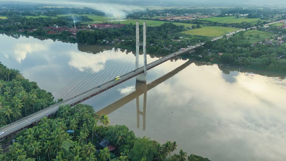 A bridge over a river with surrounding greenery and a distant industrial area.