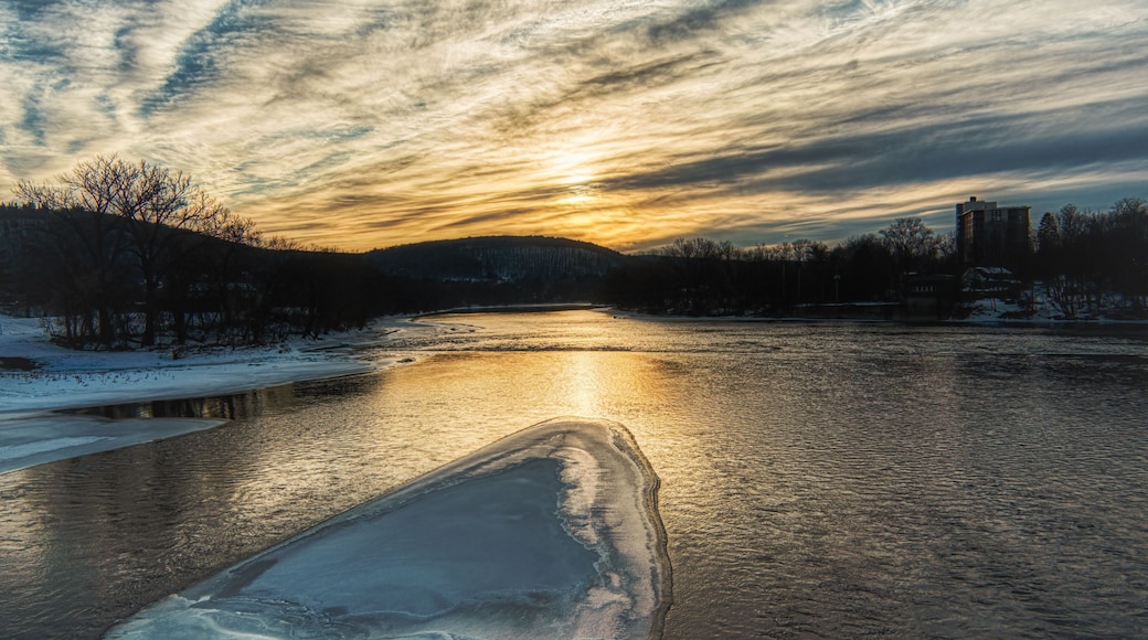 A frozen river with a partially frozen surface, surrounded by trees and buildings under a sunset sky.