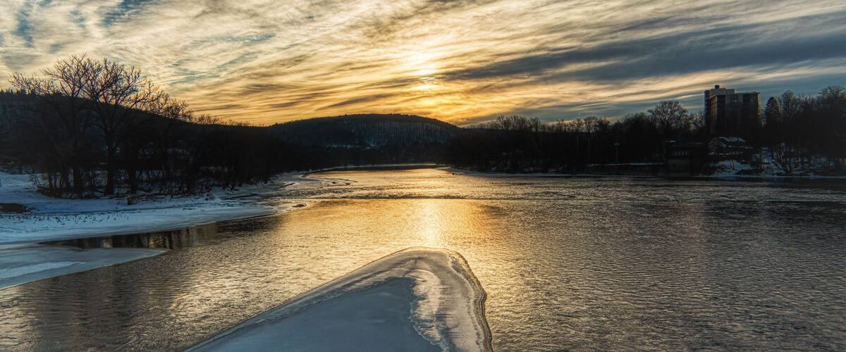 A frozen river with a partially frozen surface, surrounded by trees and buildings under a sunset sky.